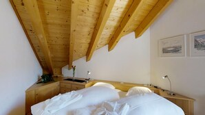 Ein Schlafzimmer mit schrägen Wänden, Holzbalken, einem Bett mit weißen Bettwäsche, einem Nachttisch mit Lampe und zwei gerahmten Bildern an der Wand.