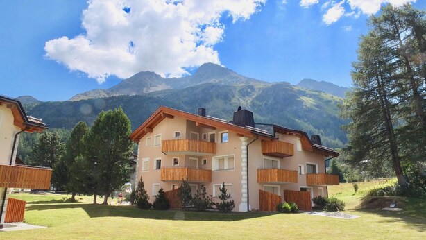Un edificio de varios pisos con balcones de madera, rodeado de un césped cuidado y árboles, con montañas al fondo.