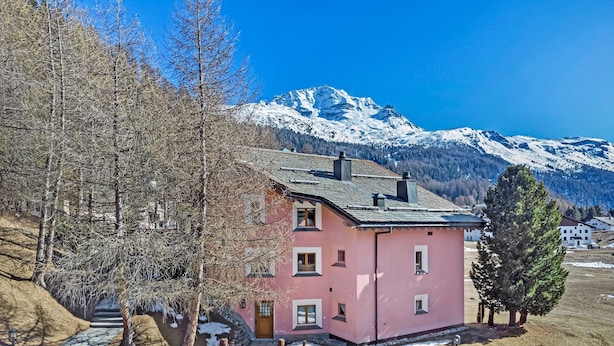 A pink house with a stone pathway leading to the entrance, surrounded by trees and snow-covered mountains in the background. - 3 ½ -room apartment on the ground floor ca. (Silvaplana-Surlej)