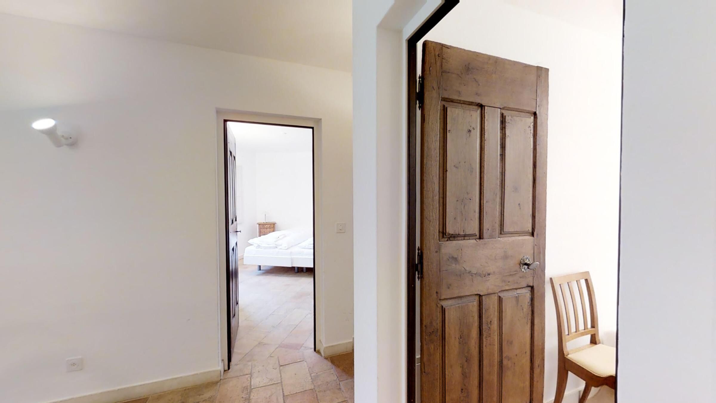A hallway with a wooden door and a chair, leading to a bedroom with a bed and a nightstand.