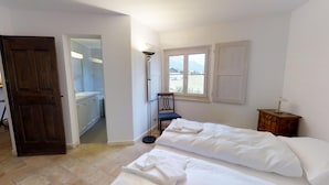 A bedroom with a bed, a chair, a desk, a window, and a door leading to a bathroom. - 3 ½ -room apartment on the ground floor ca. (Silvaplana-Surlej)