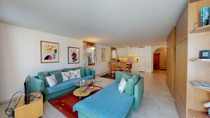 A living room with a sofa set, a coffee table, and a TV. There is a kitchen in the background with a dining table and chairs. - 2½-roomed apartment on the first floor, ca. (Silvaplana)