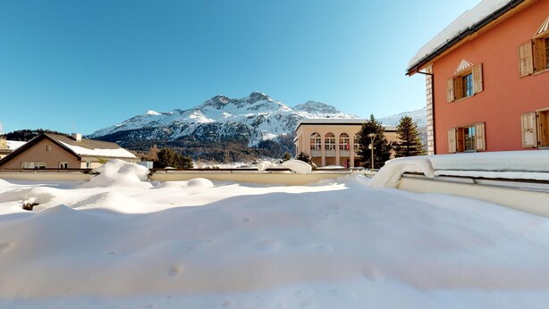 A snowy landscape with a mountain in the background, a building with arched windows, and a snow-covered roof. - 2½-roomed apartment on the first floor, ca. (Silvaplana)