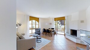 A modern living room with a sofa, chairs, a dining table, and a TV. There are large windows and a fireplace. - Modern 2½-room apartment in the first floor, about 60m2. (Maloja)