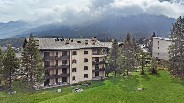 A multi-story building with balconies, surrounded by a grassy area and trees, with mountains in the background. - Modern 2½-room apartment in the first floor, about 60m2. (Maloja)