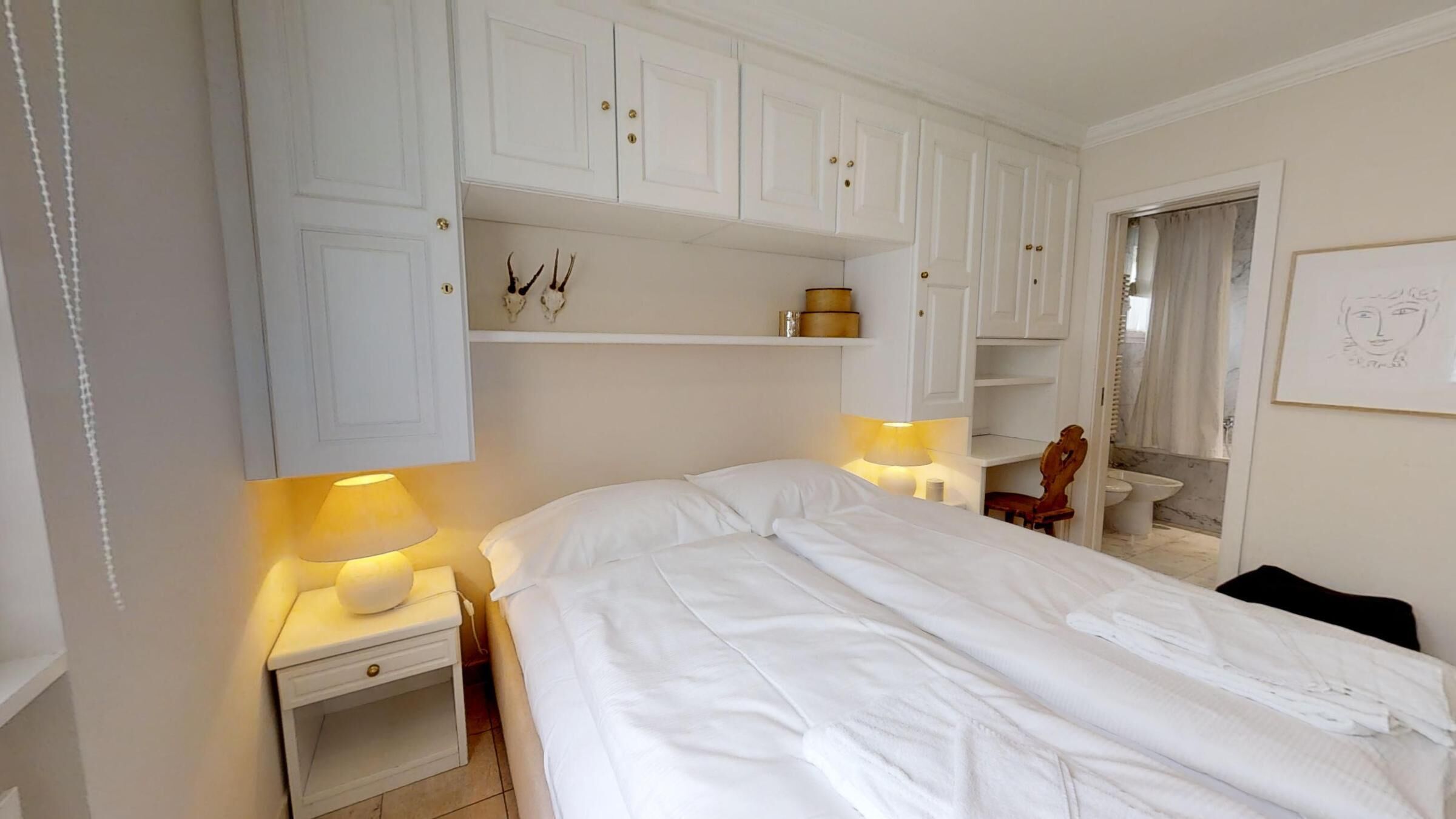 Un dormitorio con una cama, dos mesitas de noche con lámparas, una silla de madera y una pequeña mesa con una lámpara. Hay armarios blancos sobre la cama y un cuadro en la pared.