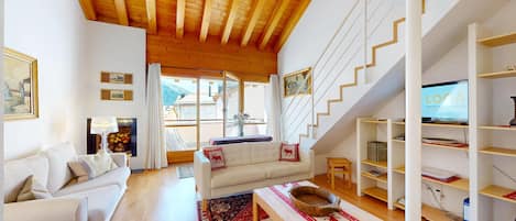 A spacious living room with a high wooden ceiling, a white staircase, and a television displaying 'LOCA'.