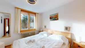 Ett sovrum med en säng i trä, ett fönster med gardiner, ett element och en tavla på väggen.