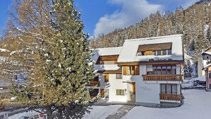 En tvåvånings chalet med en träbalkong, omgiven av snötäckta träd och en bergig bakgrund.