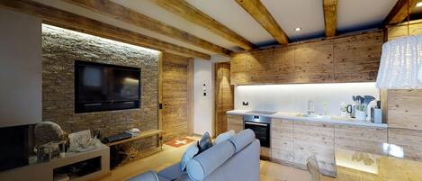 A modern living room with a stone wall, wooden beams, a flat-screen TV, a sofa, and a kitchen area with wooden cabinets and a countertop.