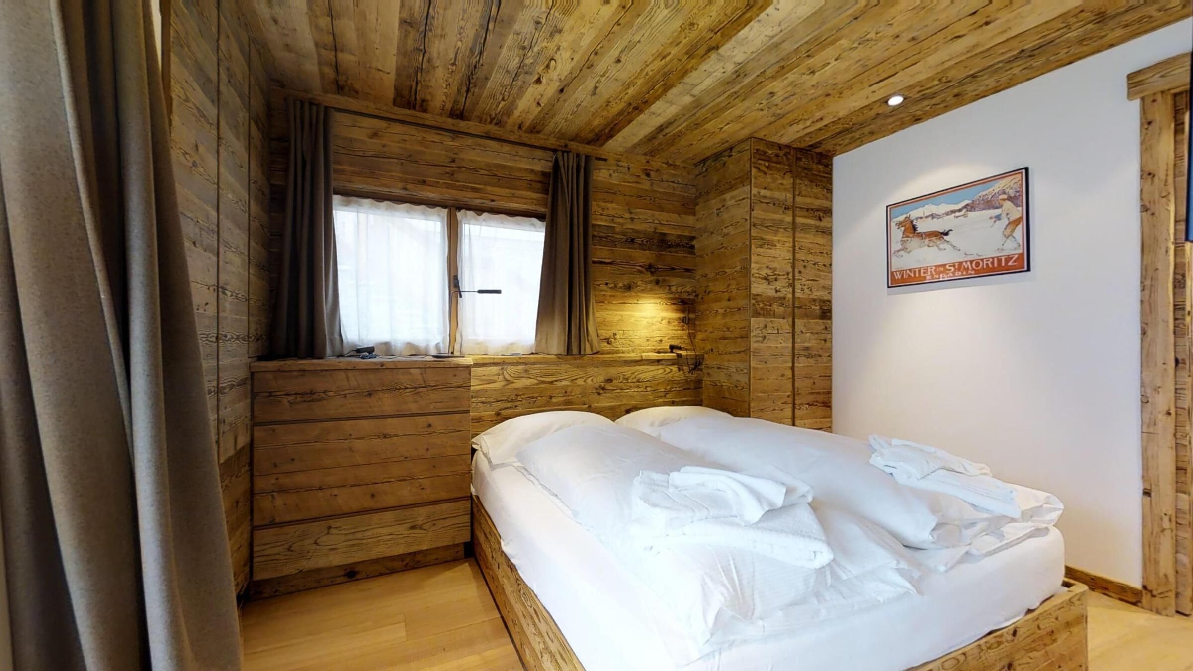 A wooden cabin room with a bed, two windows with curtains, and a poster on the wall.