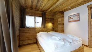 A wooden cabin room with a bed, two windows with curtains, and a poster on the wall.