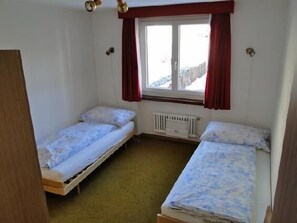 A small room with two single beds, a window with red curtains, and a radiator.