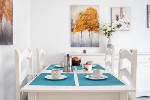 Ruokapöytä, jossa on valkoiset tuolit, kahvipannu ja hedelmäkori.