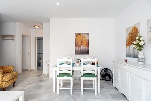 Un coin repas avec une table blanche, deux chaises, un ventilateur et un meuble.