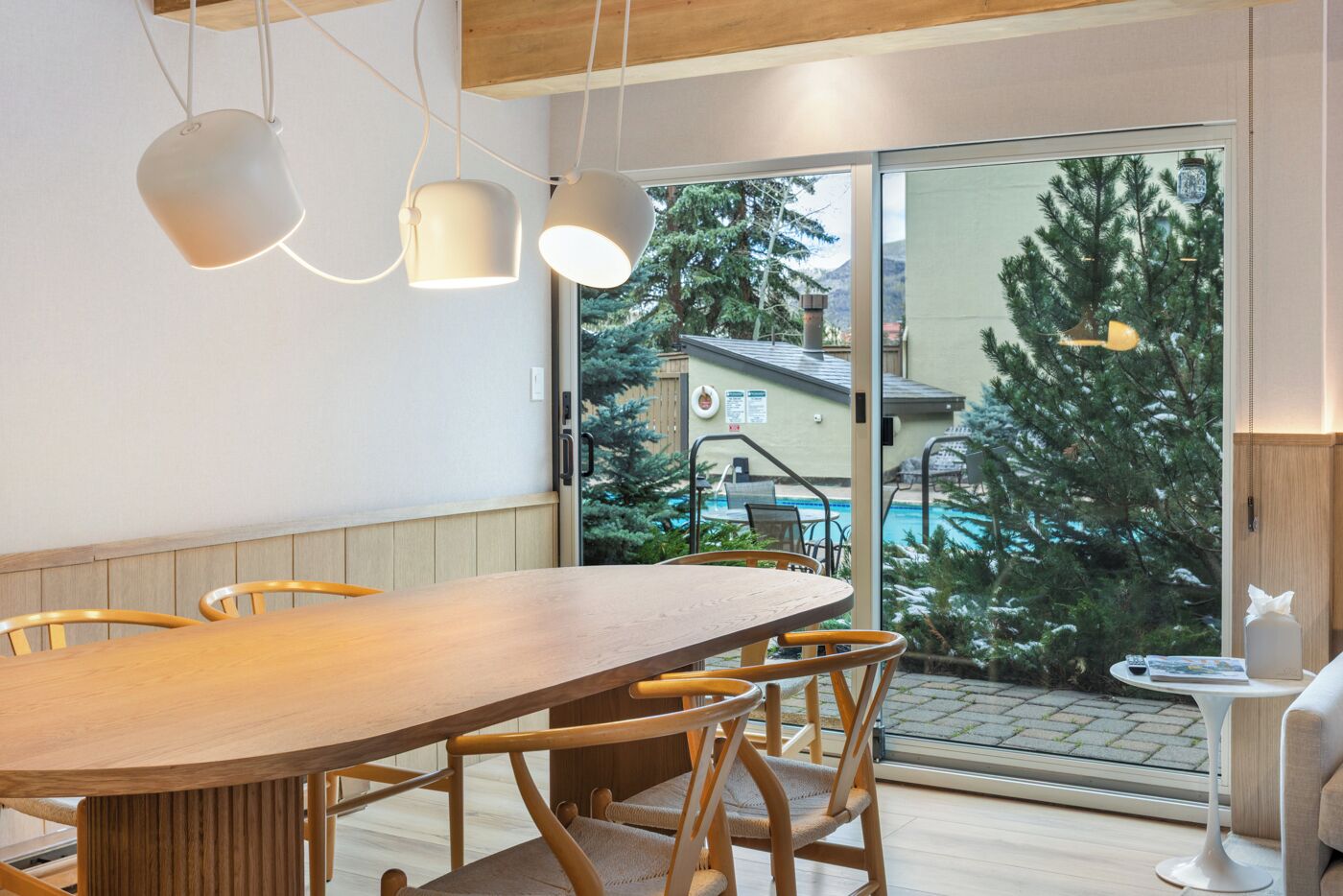 Ein moderner Essbereich mit Holztisch und -stühlen, Pendelleuchten und Blick auf Pool und Bäume durch große Glasschwingtüren.