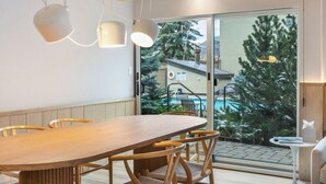 A modern dining area with a wooden table and chairs, pendant lights, and a view of a pool and trees through large glass doors.