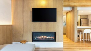 A modern living room with a flat-screen TV mounted on a wooden wall, a fireplace below, and a kitchen visible through an open doorway.