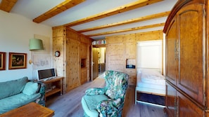 A cozy room with wooden walls and ceiling, a sofa, a floral armchair, a bed, and a desk with a lamp.