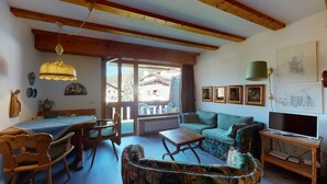 A cozy living room with a sofa, a wooden coffee table, and a TV stand. There is a dining area with a table and chairs, and a large window offering a view of the outside.
