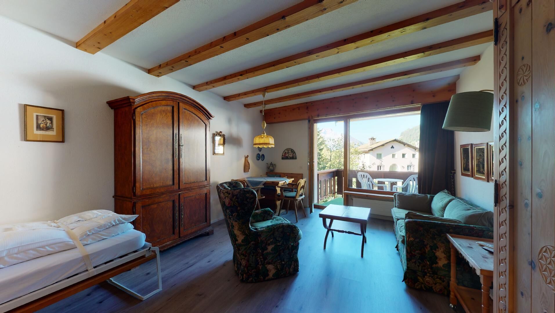 A cozy room with a bed, a wooden cabinet, a floral armchair, a sofa, and a dining table.