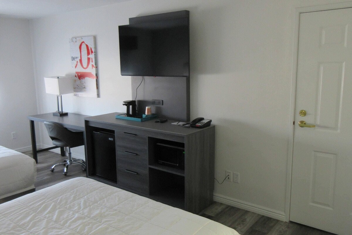 A hotel room with a bed, a desk with a chair, a flat-screen TV, and a door.