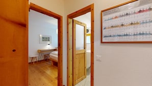 Um quarto com porta de madeira, uma cama, uma escrivaninha e um quadro fixado na parede.