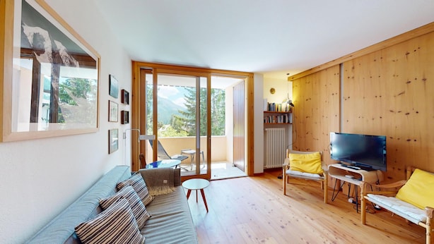 Uma sala de estar moderna com um sofá, cadeiras, uma TV e uma varanda com vista para árvores e montanhas.