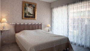 A bedroom with a bed, two bedside lamps, a framed painting on the wall, and sheer curtains on the window. - Le Lavandou, One-bedroom duplex to rent for 4 people, glimpse of the sea, near city-center and... (LE LAVANDOU)