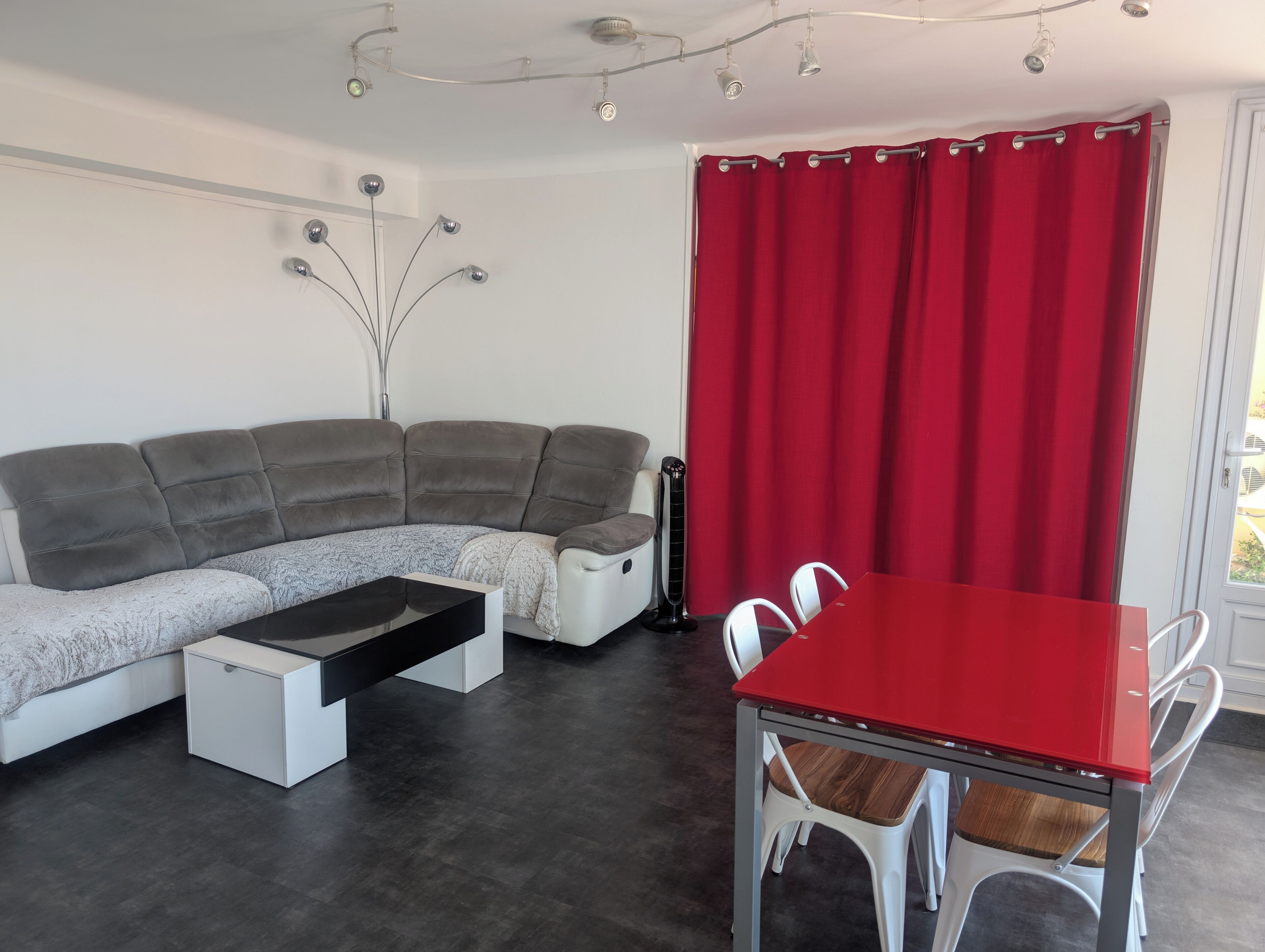 Ein modernes Wohnzimmer mit einem grauen Ecksofa, einem kleinen weißen Tisch und einem roten Esstisch mit weißen Stühlen.