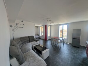 A modern living room with a grey sectional sofa, a coffee table, and a dining area with a table and chairs. There is a large window providing natural light and a view of the outside. - 58m2 flat in ST Raphael-600 metres from the beach and train station-free parking (Saint-Raphaël)