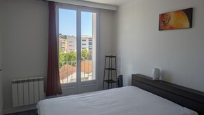 A hotel room with a bed, a radiator, a window with curtains, a small table with a vase, and a painting on the wall. - 58m2 flat in ST Raphael-600 metres from the beach and train station-free parking (Saint-Raphaël)