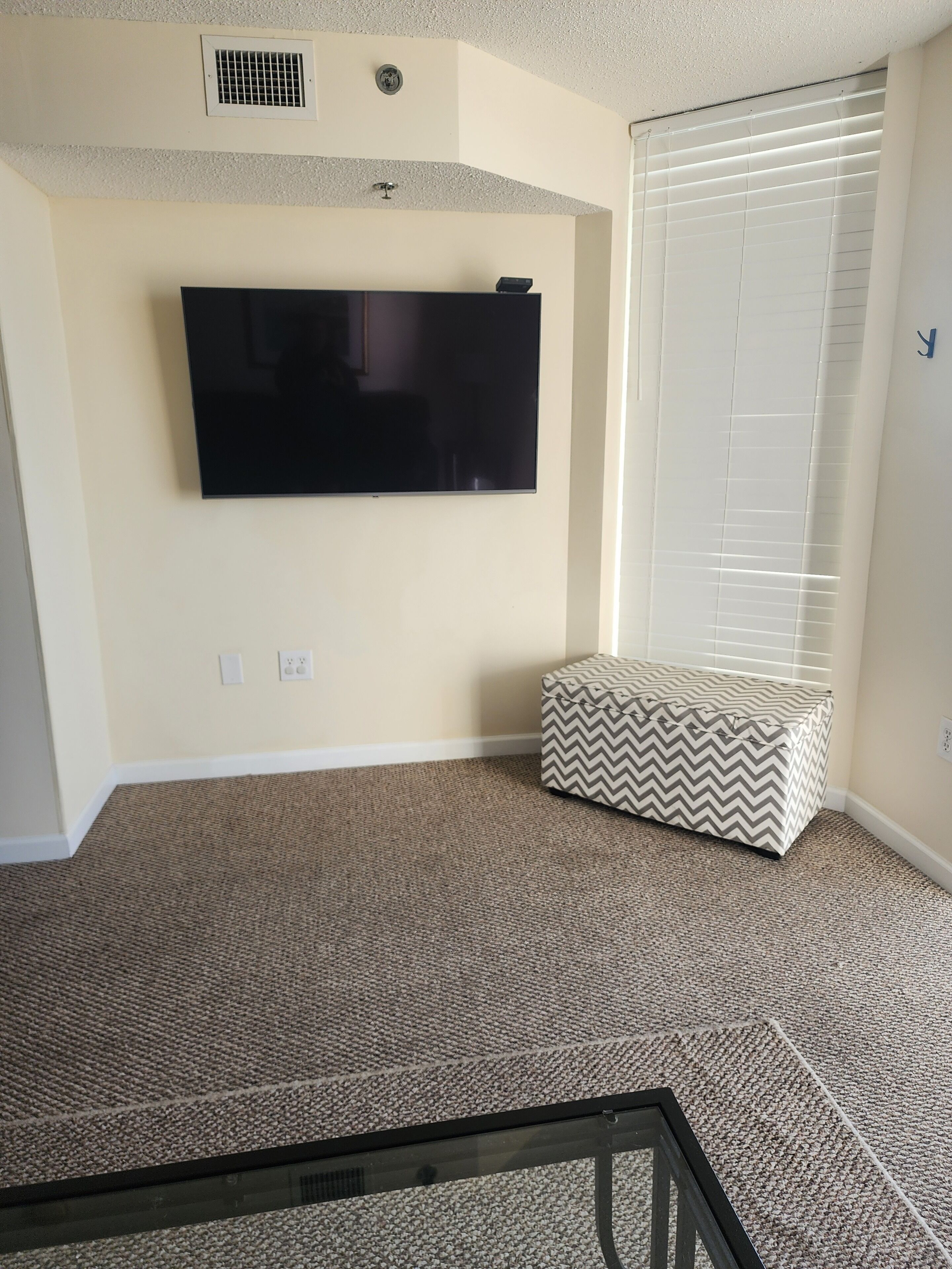 A living room with a flat-screen TV mounted on the wall, a patterned ottoman, a carpeted floor, and a window with blinds.