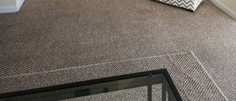 A living room with a flat-screen TV mounted on the wall, a patterned ottoman, a carpeted floor, and a window with blinds.