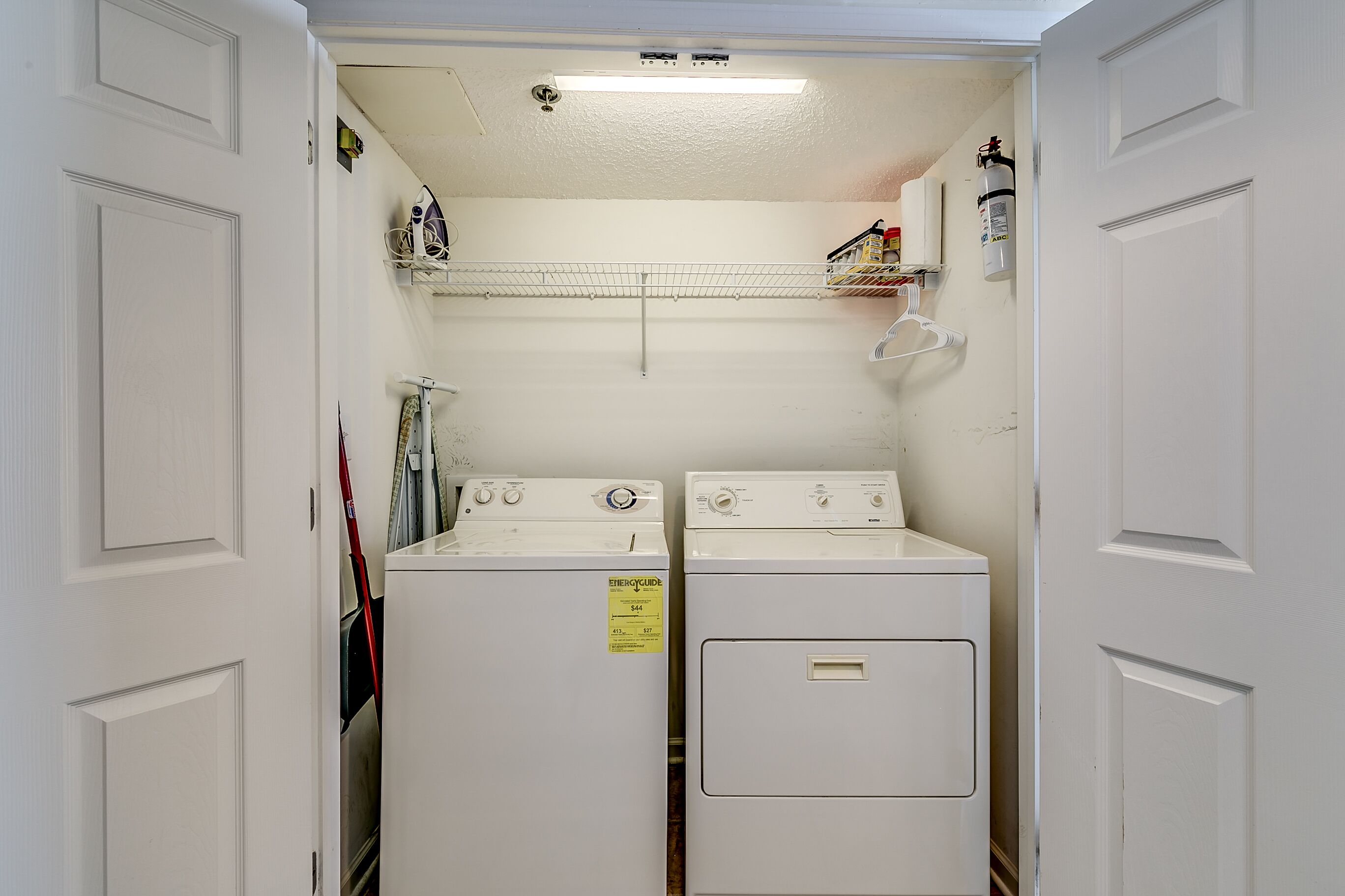Une buanderie équipée de deux machines à laver et deux sèche-linge superposés, d’une étagère contenant des produits de nettoyage et d’un luminaire au plafond.