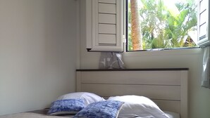 A bedroom with a bed, a window with a view of greenery, and a white air conditioning unit. - DISCOVER MY CHARMING BUNGALOW WITH POOL (Le Gosier)