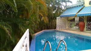 A swimming pool with a white deck and metal handrails, surrounded by lush greenery and a blue-roofed structure. - DISCOVER MY CHARMING BUNGALOW WITH POOL (Le Gosier)