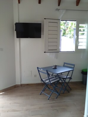 A room with a table and chairs, a window with shutters, and a mounted television. - DISCOVER MY CHARMING BUNGALOW WITH POOL (Le Gosier)