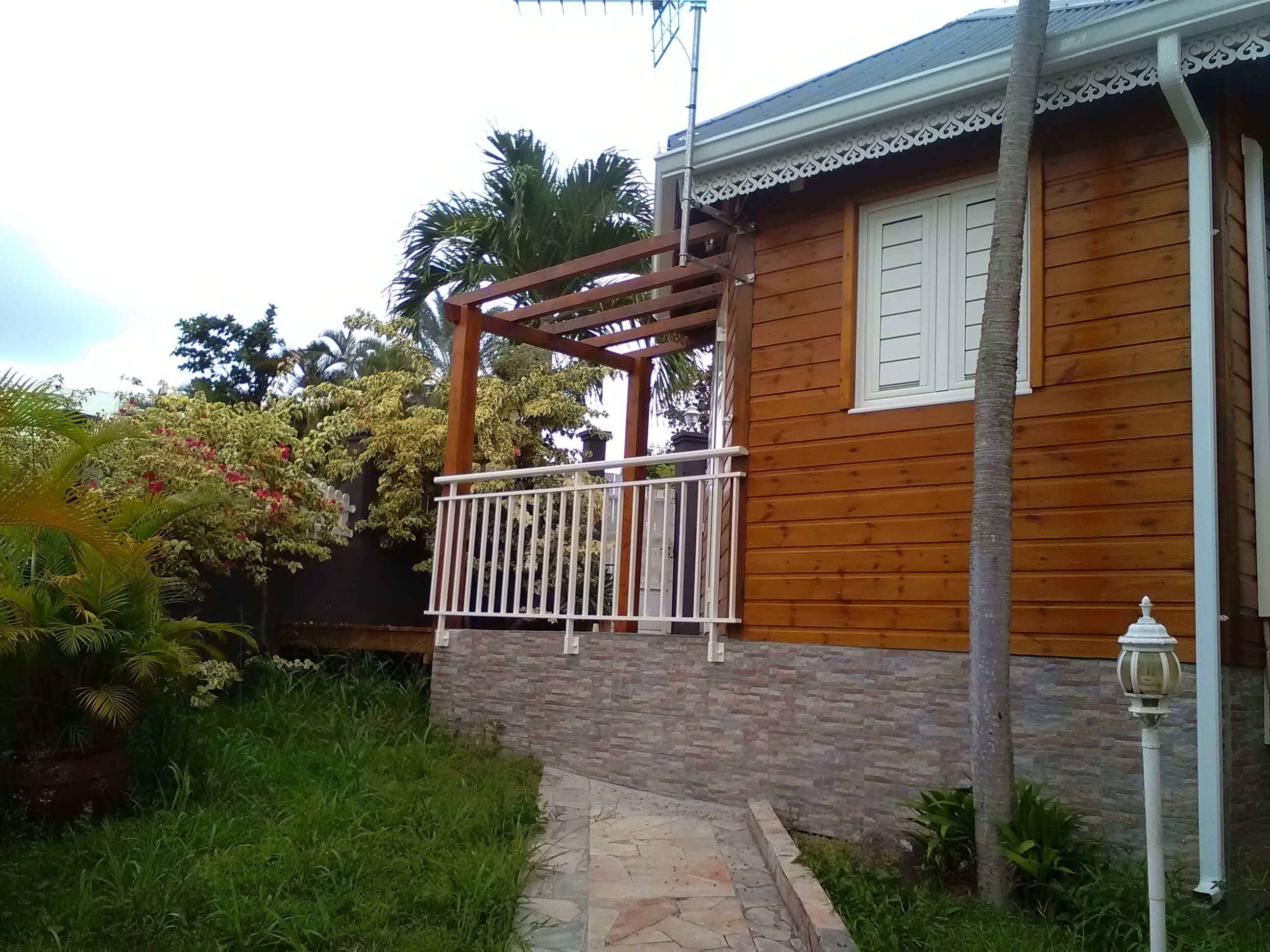 Une maison en bois avec un balcon, un chemin de pierre et un jardin où poussent différentes espèces de plantes.