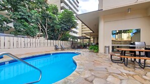 Une piscine avec des carreaux de pierre, des chaises longues, un barbecue et un espace salon couvert offrant une vue sur le bâtiment.