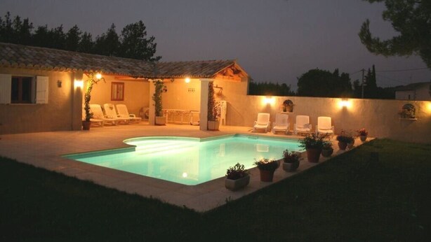 A swimming pool with lounge chairs and potted plants, surrounded by a building with a tiled roof and exterior lighting.