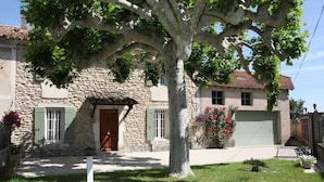 A stone building with a large tree in front, a door, and a small garden with flowers.