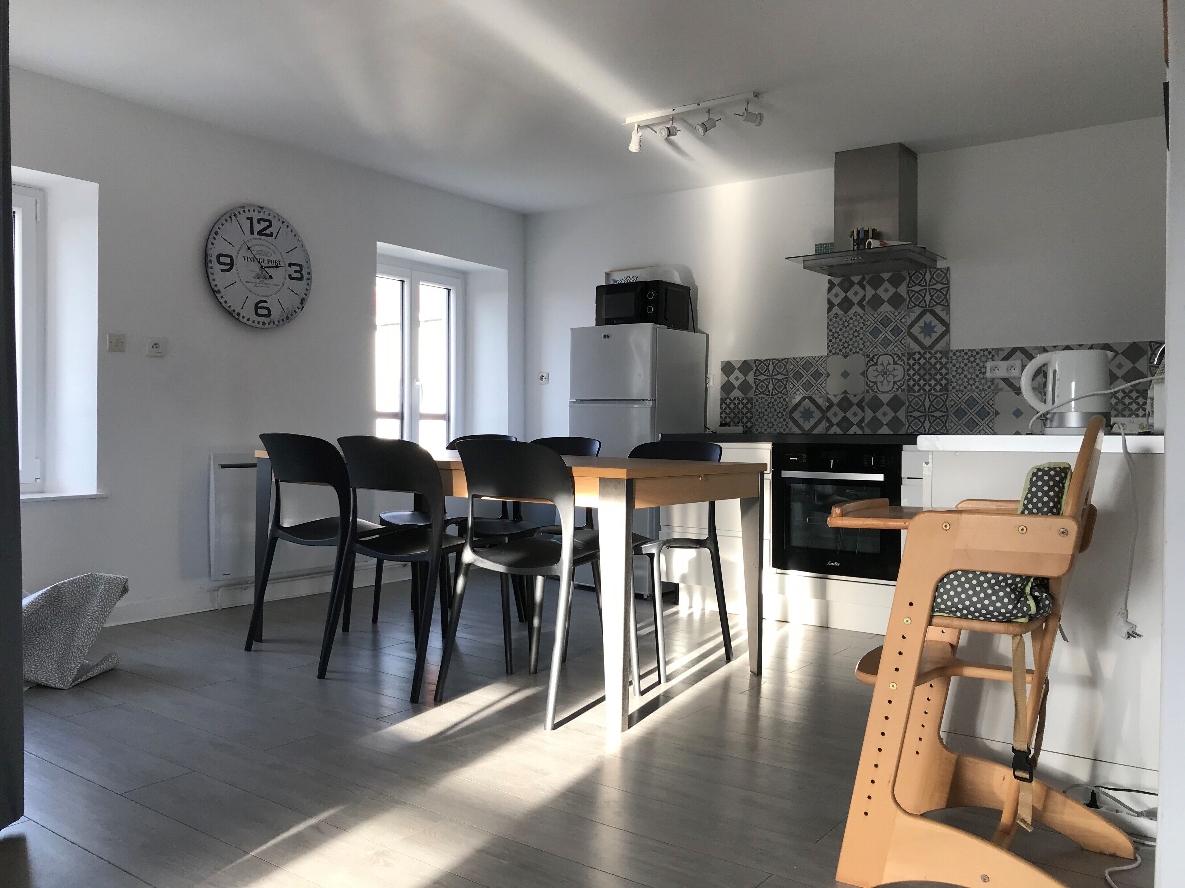 A modern kitchen with a dining area, featuring a refrigerator, oven, and a clock on the wall.