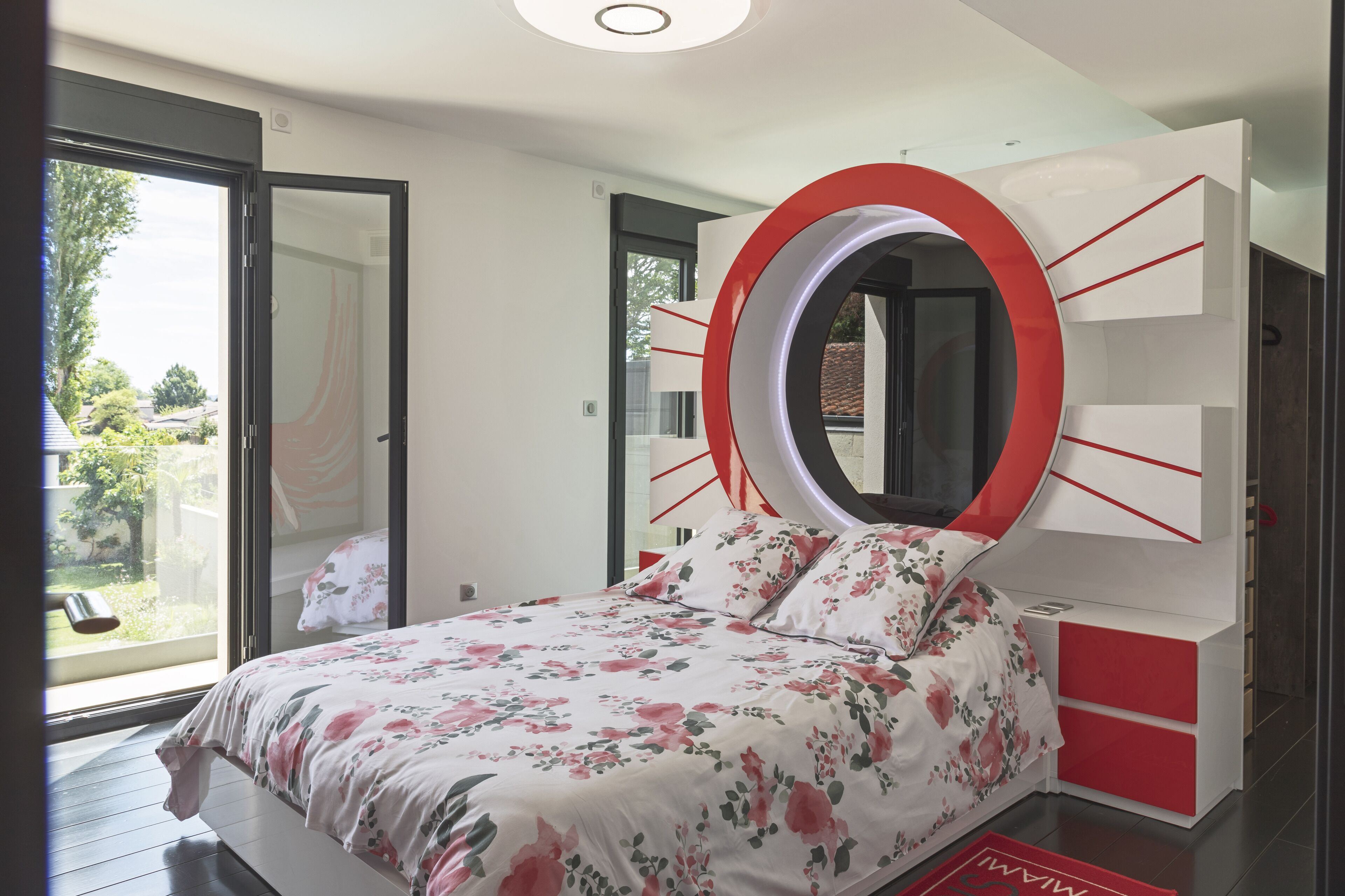 A bedroom with a bed, a circular mirror, and a red and white dresser.