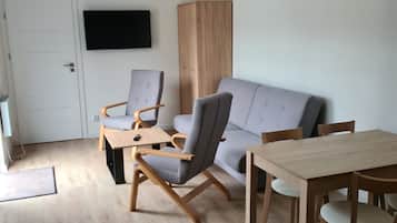 A modern living room with a grey sofa, wooden coffee table, and matching chairs. A flat-screen TV is mounted on the wall, and a wooden cabinet is visible.