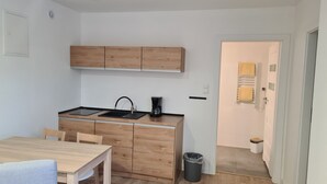 Cocina compacta con armarios de madera, un lavamanos negro y una cafetera. Hay un pequeño cubo de basura y una puerta que da a un baño con un perchero.