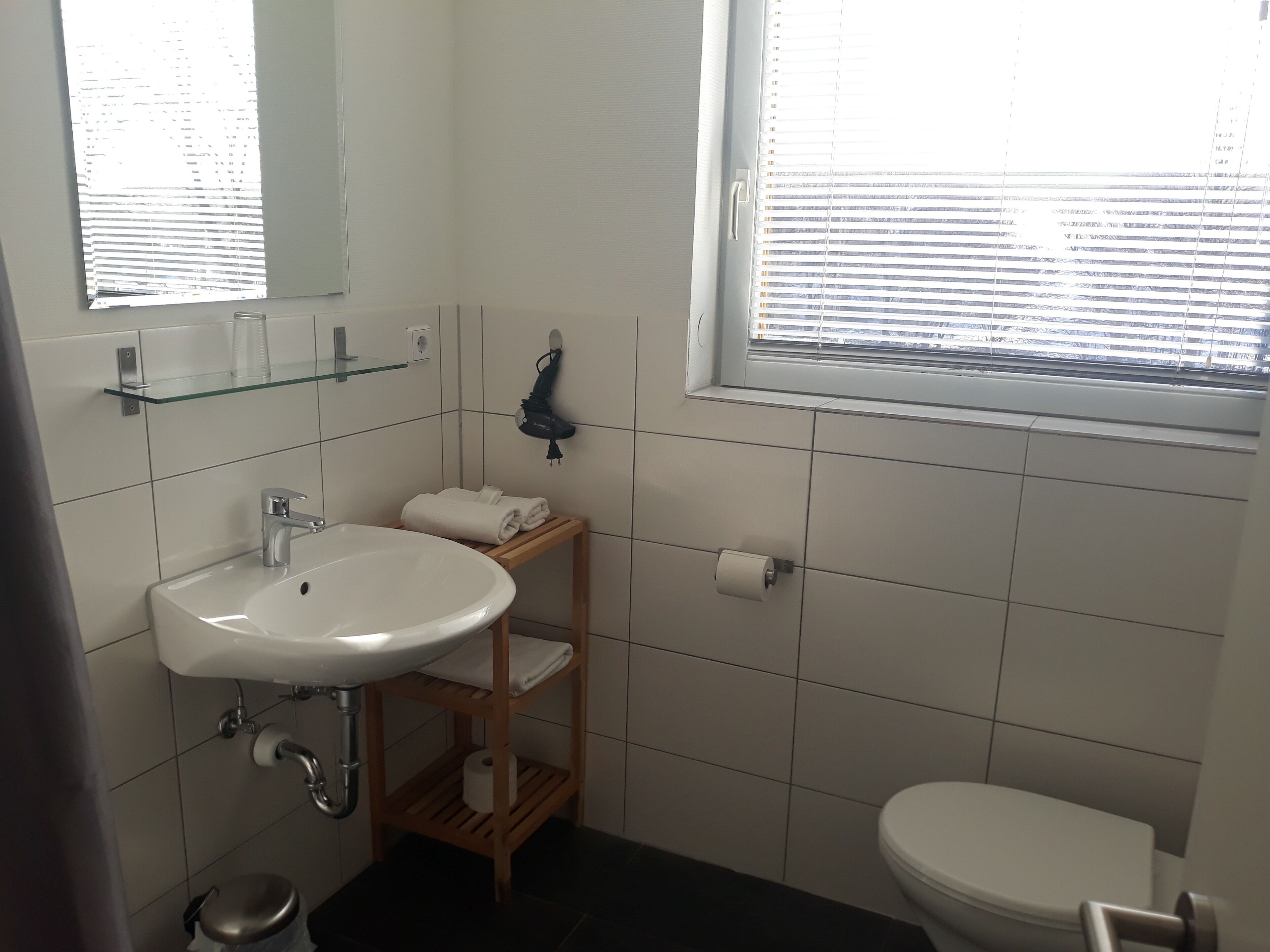 A bathroom with a white sink, mirror, and toilet. There is a towel rack and a glass shelf above the sink.