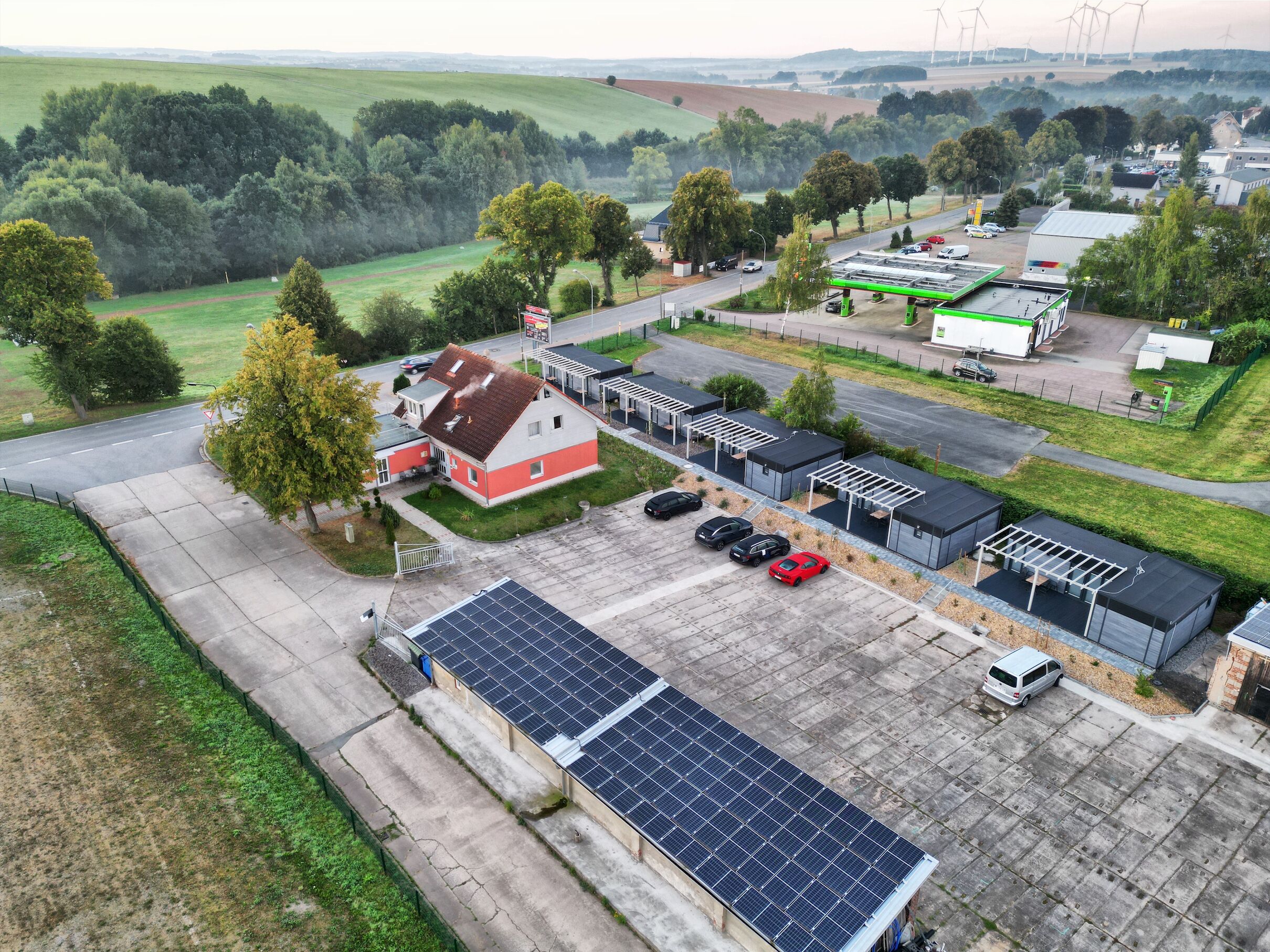 Vista aérea de uma zona rural com uma grande instalação de painéis solares, um edifício vermelho e branco e vários carros estacionados.