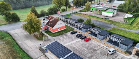 An aerial view of a rural area with a large solar panel installation, a red and white building, and several parked cars.
