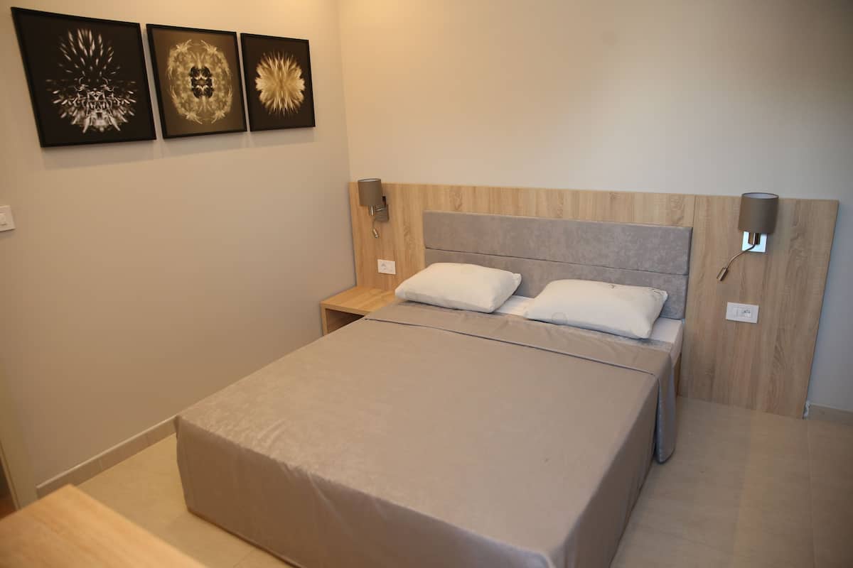 A neatly made bed with a grey bedspread and two pillows, a wooden headboard, a nightstand with a lamp, and three wall art pieces above the bed.